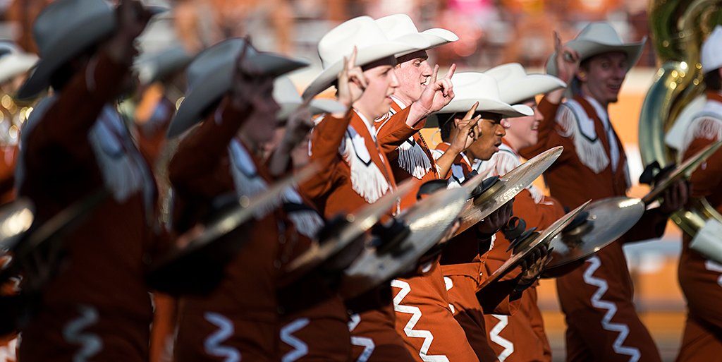 UT Band Cymbal Section marching in burnt orange uniforms with hands raised showing the hookem horns sign 