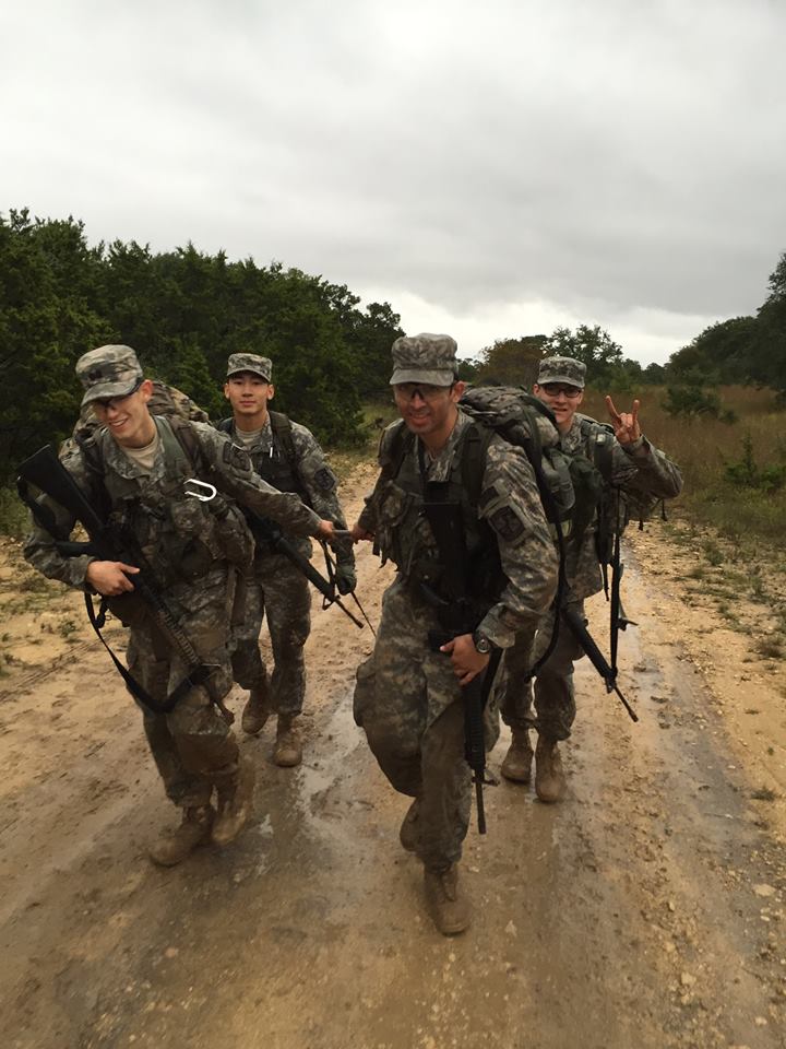 Four UT ROTC students shown walking up a muddy path with one student showing the hookem horns sign with his hand.
