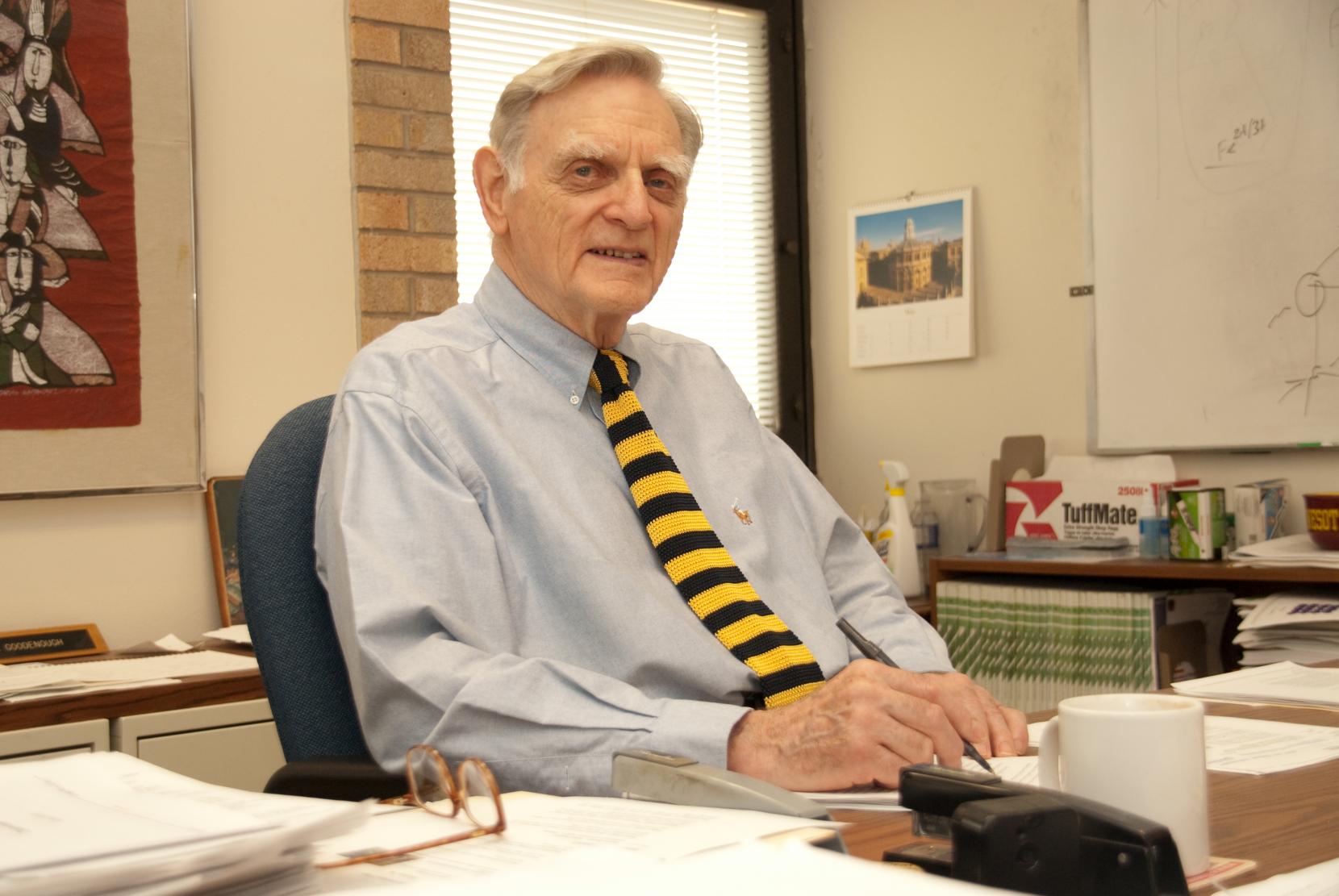 John Goodenough poses at his desk holding a pen and looking at the camera smiling