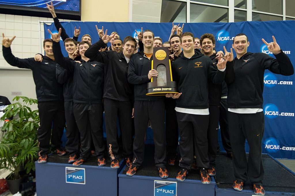Swimming and Diving team students stand on podium holding a trophy with students showing the hookem sign with their hands