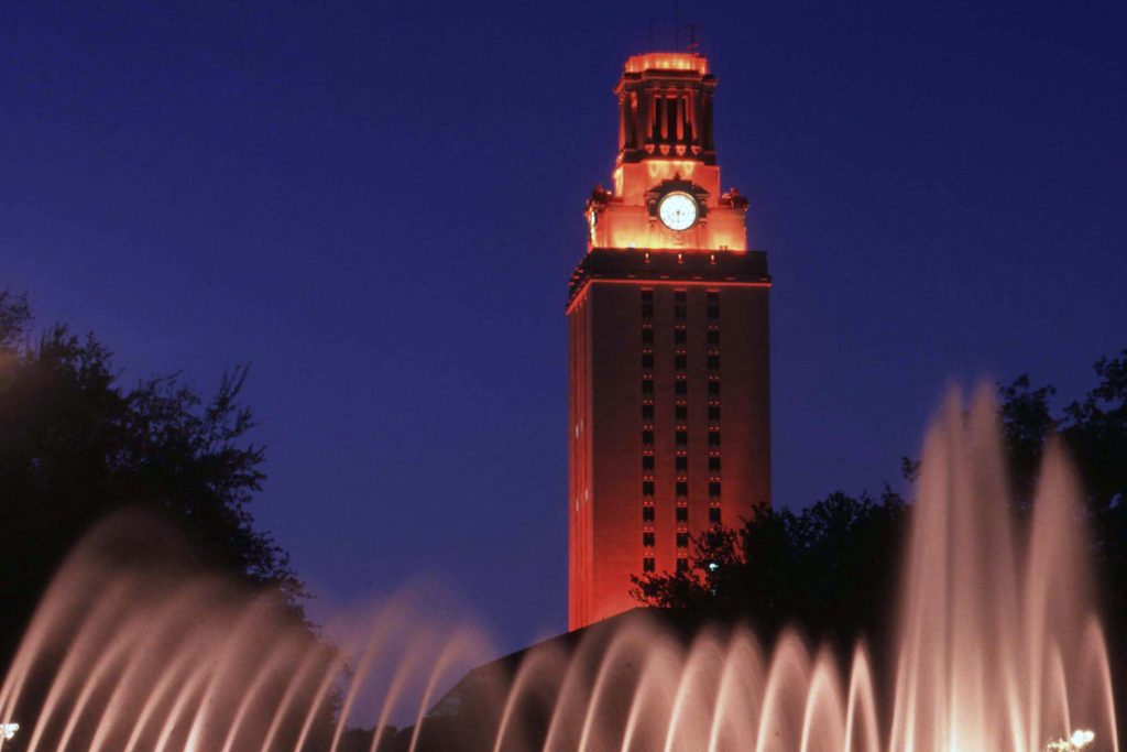 UT Tower glowing orange against a dark blue sky with fountains seen in the foreground 