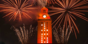 The UT Tower shines with burnt orange lights and the number “16” displayed on its sides and fireworks in the background.