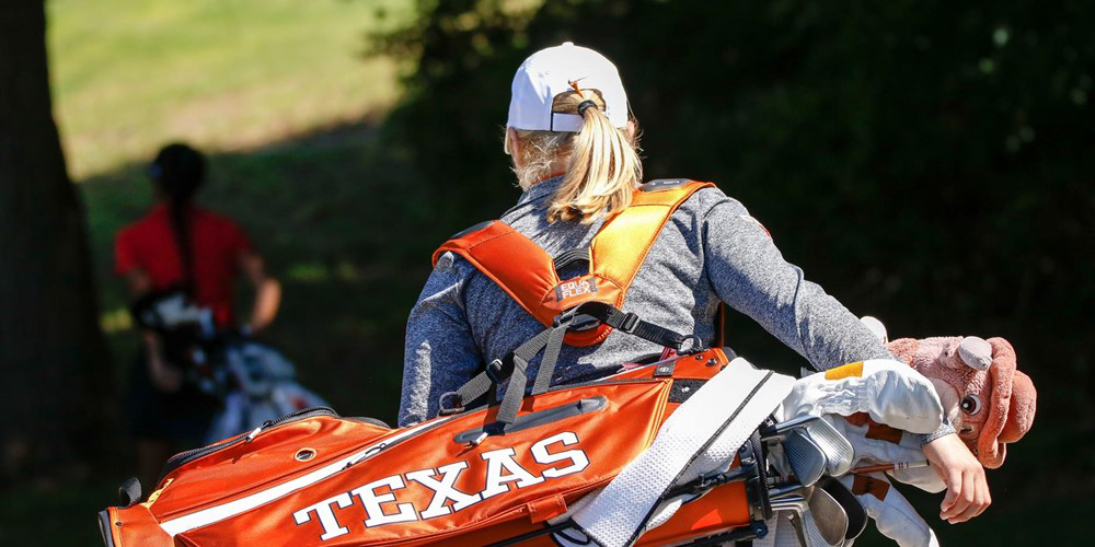 UT Women's golf student shown from behind carrying an orange golf bag with the word Texas on it