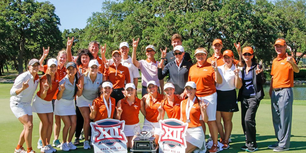 UT Golf team pictured on the golf course posing with hands showing the hookem sign with a large trophy next to signs that read XII Championship 2017 Big 12 Women's Golf Champion