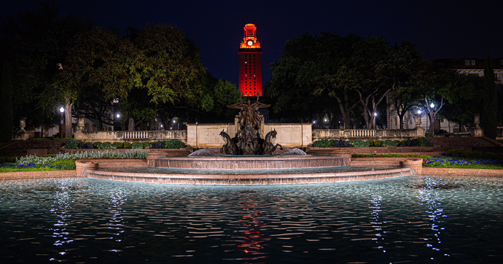 The UT Tower shines with burnt orange lights at night with Littlefield Fountain in the foreground