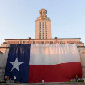 A Texas flag hangs from the front of the Main Building as the UT Tower stands in the background.