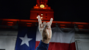 A hand making the “Hook Em, Horns” signs with the UT Tower and Texas flag in the background.