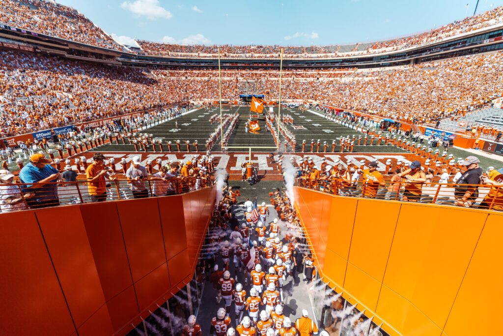 View of DKR-TMS shows fans filling the stands as the football team enters the field through a tunnel. 