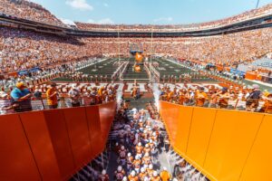 View of DKR-TMS shows fans filling the stands as the football team enters the field through a tunnel.