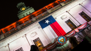 A Texas flag hangs from the front of the Main Building as the UT Tower stands in the background at night.