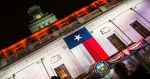 A Texas flag hangs from the front of the Main Building as the UT Tower stands in the background at night.