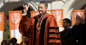 President Jay Hartzell wears academic regalia and poses for a picture with his “Hook Em Horns” hand sign up.