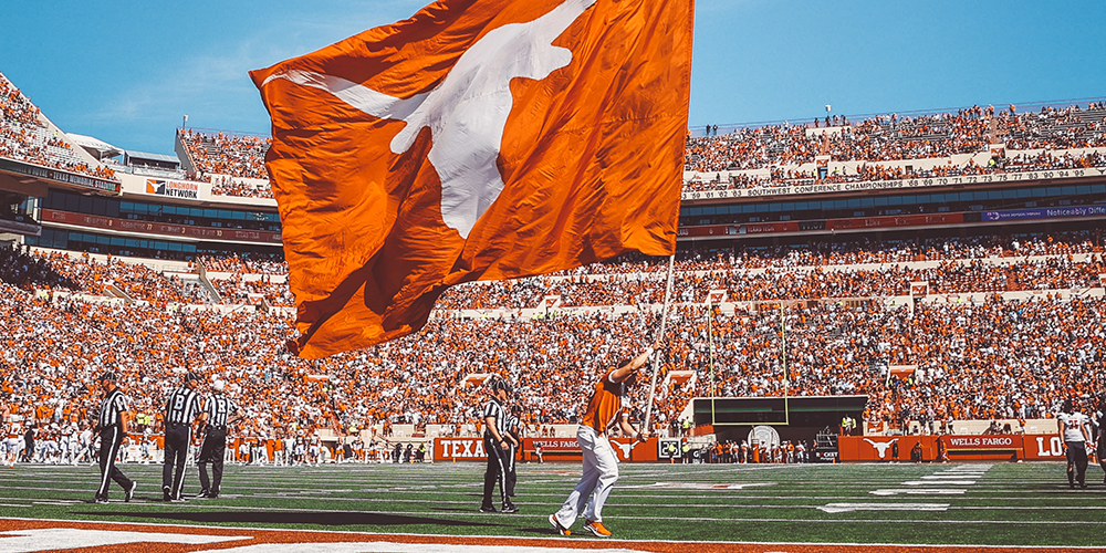 A man runs across the field of the Texas Football stadium waving a giant flag with the Longhorn silhouette as fans pack the stands in the background. 